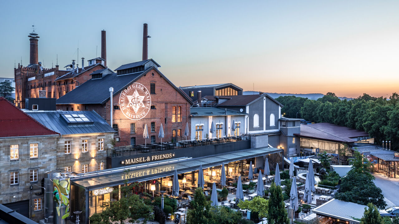 A historic brewery with outdoor seating and terraces under parasols, illuminated at dusk.