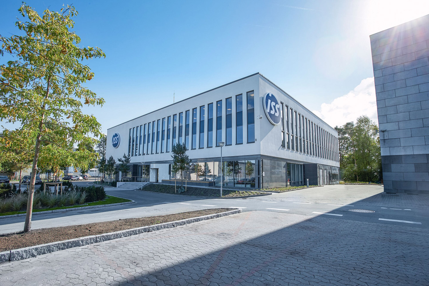 A modern, rectangular office building with large windows and the "ISS" logo on the facade, surrounded by trees and a paved forecourt, under a clear blue sky.