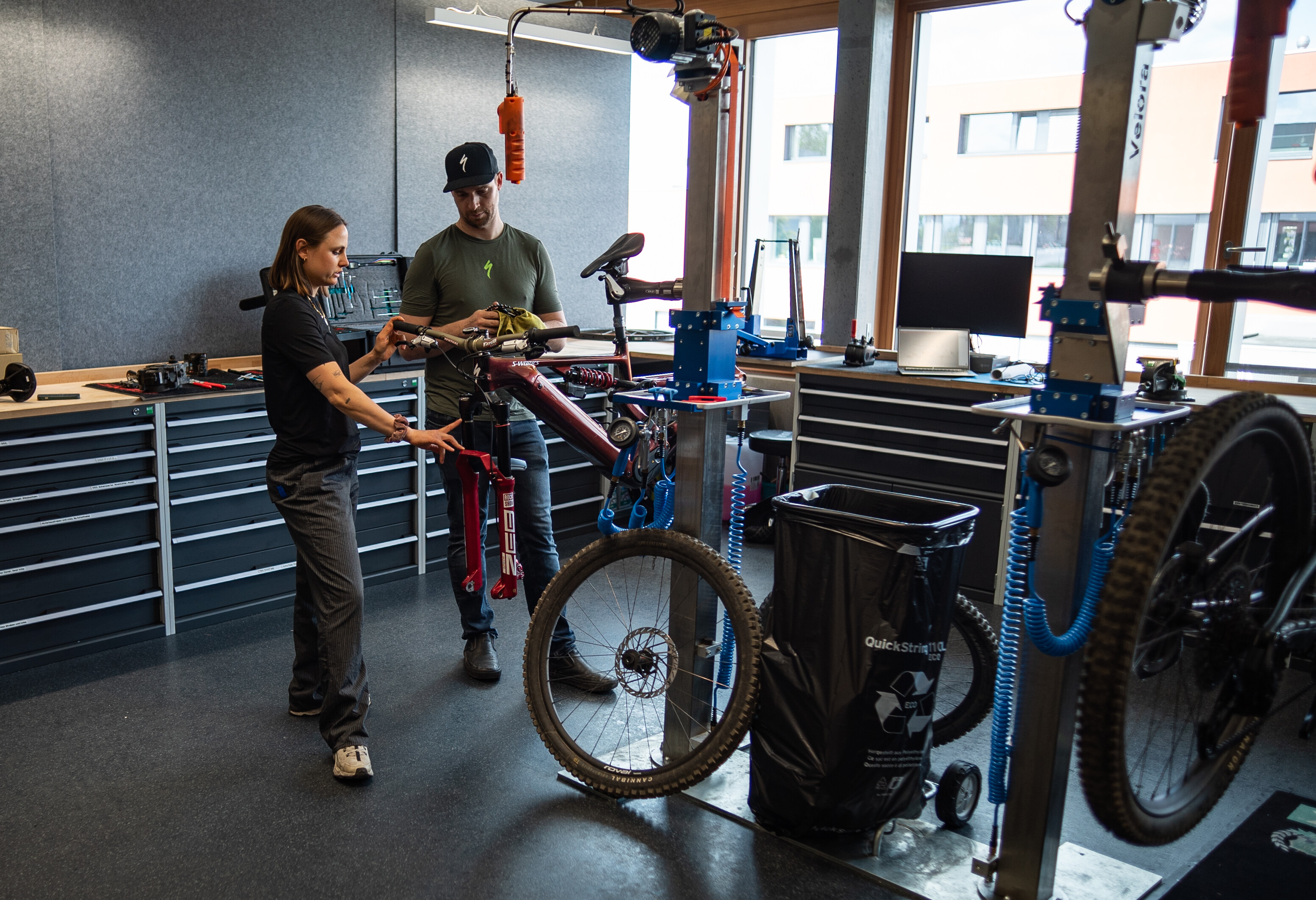 Sofia Wiedenroth is standing with a bicycle technician in a workshop next to an assembled bike. Sofia is pointing to a component while the bicycle technician stands next to her holding something in his hands.