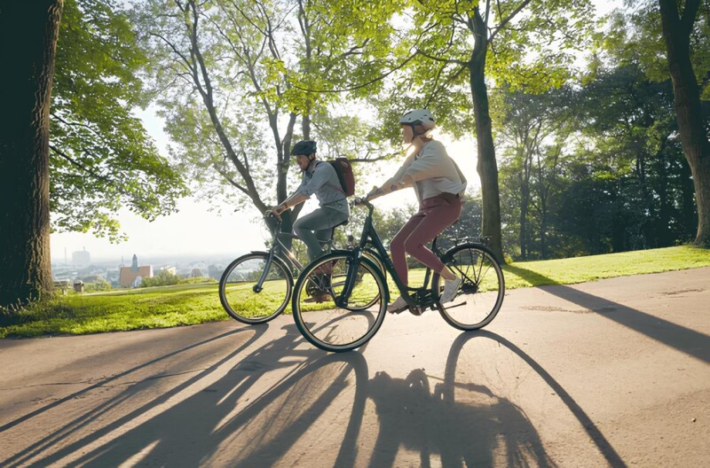Two people are riding bikes on a path through a park in sunny weather. The long shadows of the trees and cyclists stretch across the ground, and trees and a view of the city are visible in the background.