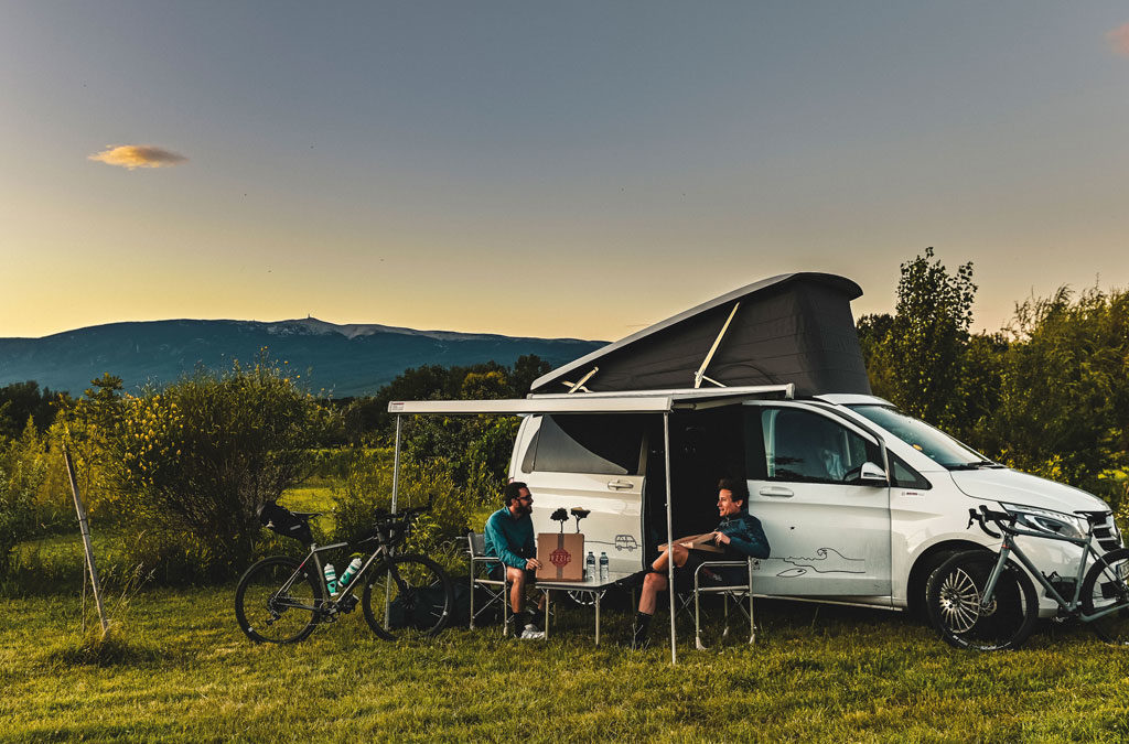 Two people are sitting on camping chairs in front of a white camper van, chatting while their bikes are parked next to the van. Trees and mountains can be seen in the background, with the evening twilight providing atmospheric lighting.