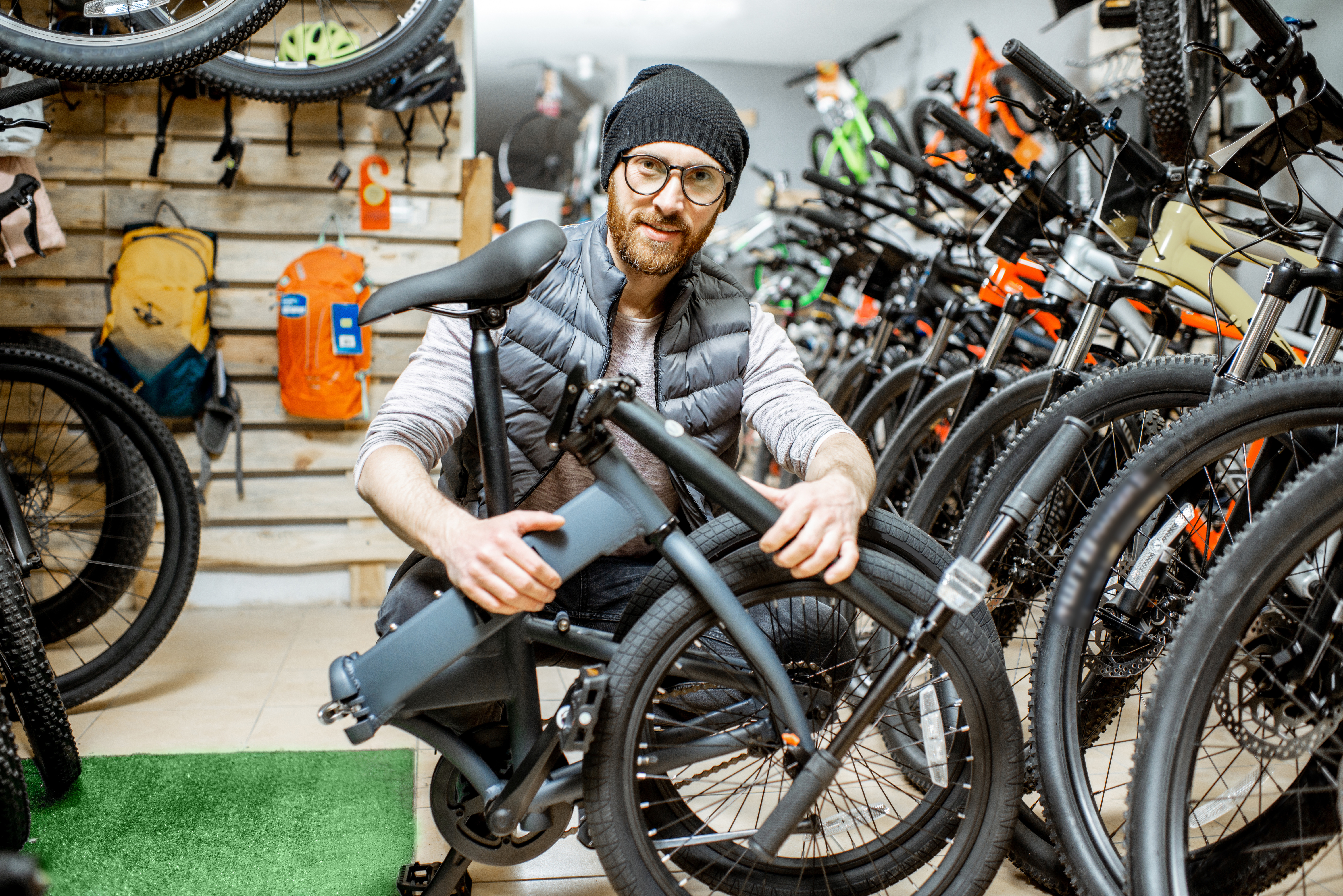 A man wearing a cap and glasses is kneeling in a bicycle shop, holding a folded folding bike.