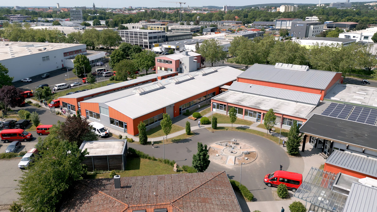 Aerial view of a building complex with flat roofs, surrounded by trees and parked red delivery trucks. In the foreground is a round square with a fountain.