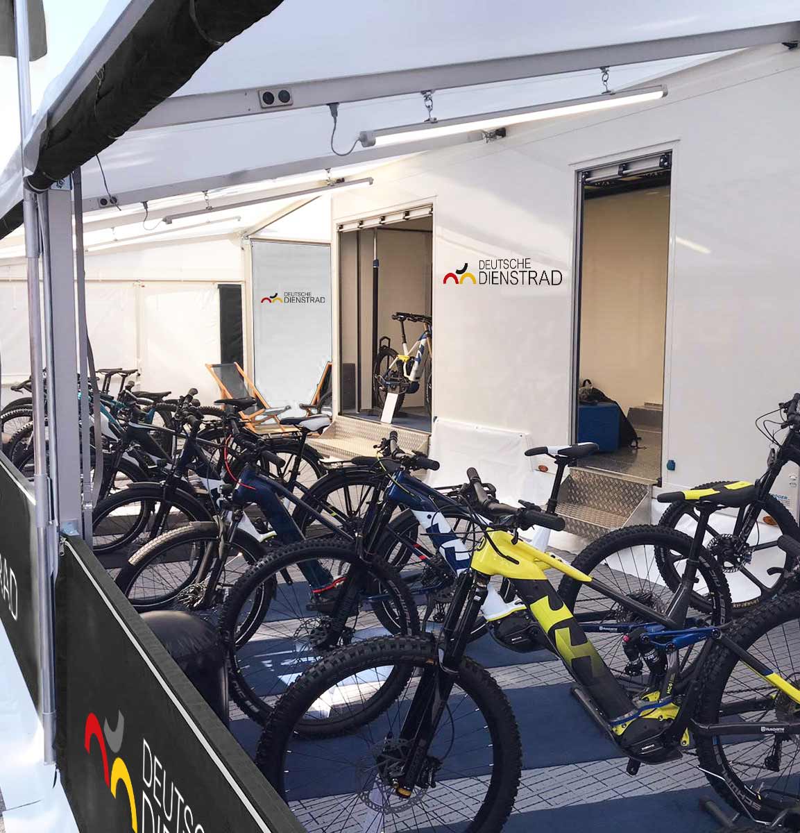 The image shows several bikes displayed in a mobile exhibition stand belonging to "Deutsche Gesellschaft für company bikes." The stand is constructed from white tarpaulins, with the company's logos on the walls. The bikes are neatly arranged and ready for presentation. Two chairs are also visible, creating a relaxed atmosphere.