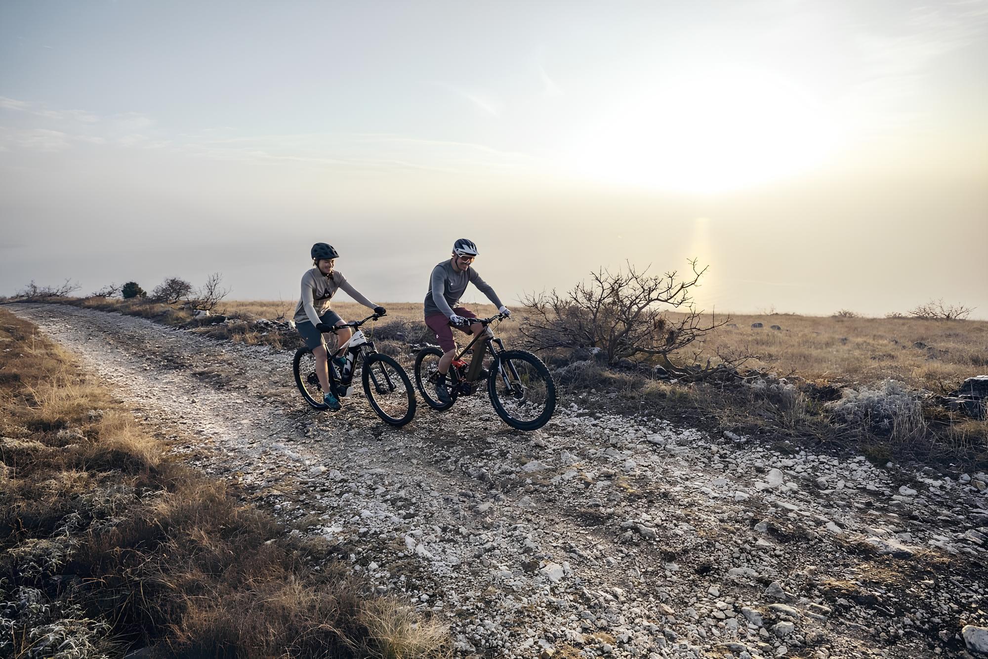 Two people ride mountain bikes on a rocky path through a barren landscape in the sunlight.