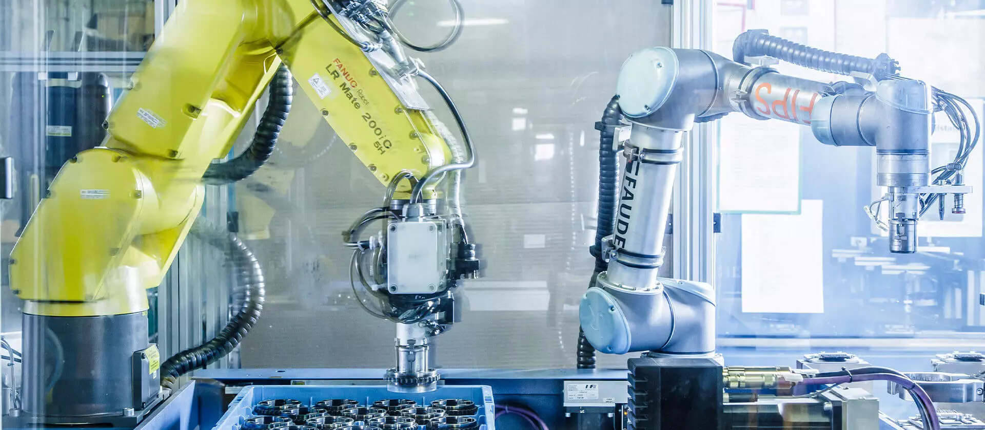 Two industrial robotic arms, one yellow and one silver, operate in an automated production environment. They move components with precise movements within a glass protective enclosure.