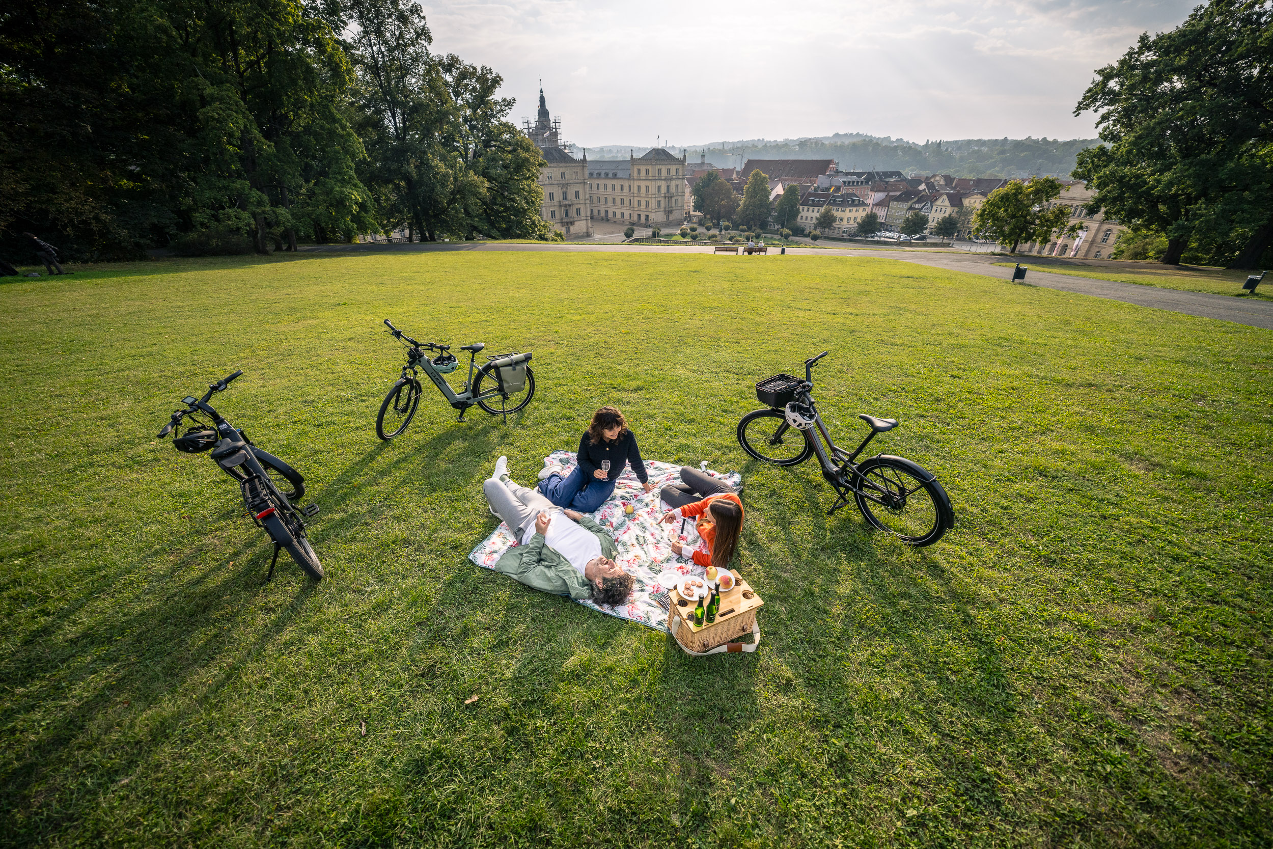 Three people are having a picnic on a lawn overlooking an old town, surrounded by e-bikes.