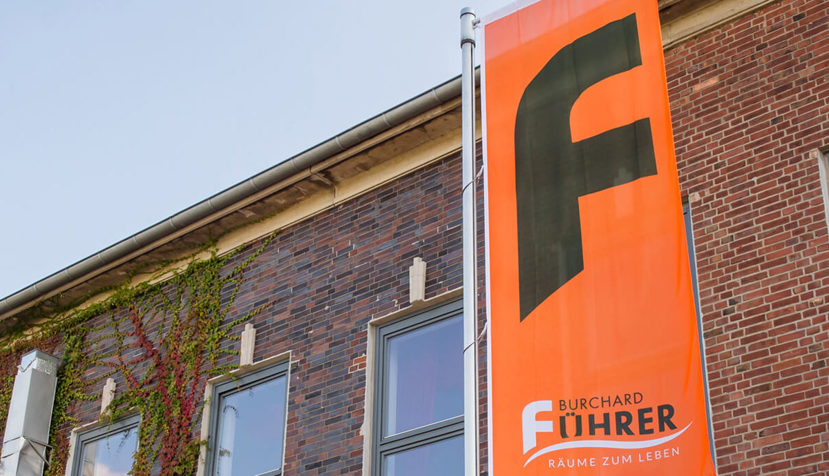 Close-up of an orange flag with a large black "F" and the text "Burchard Führer - Räume zum Leben" (Burchard Führer - Rooms to Live In) in front of a brick building covered in ivy. The sky is clear.