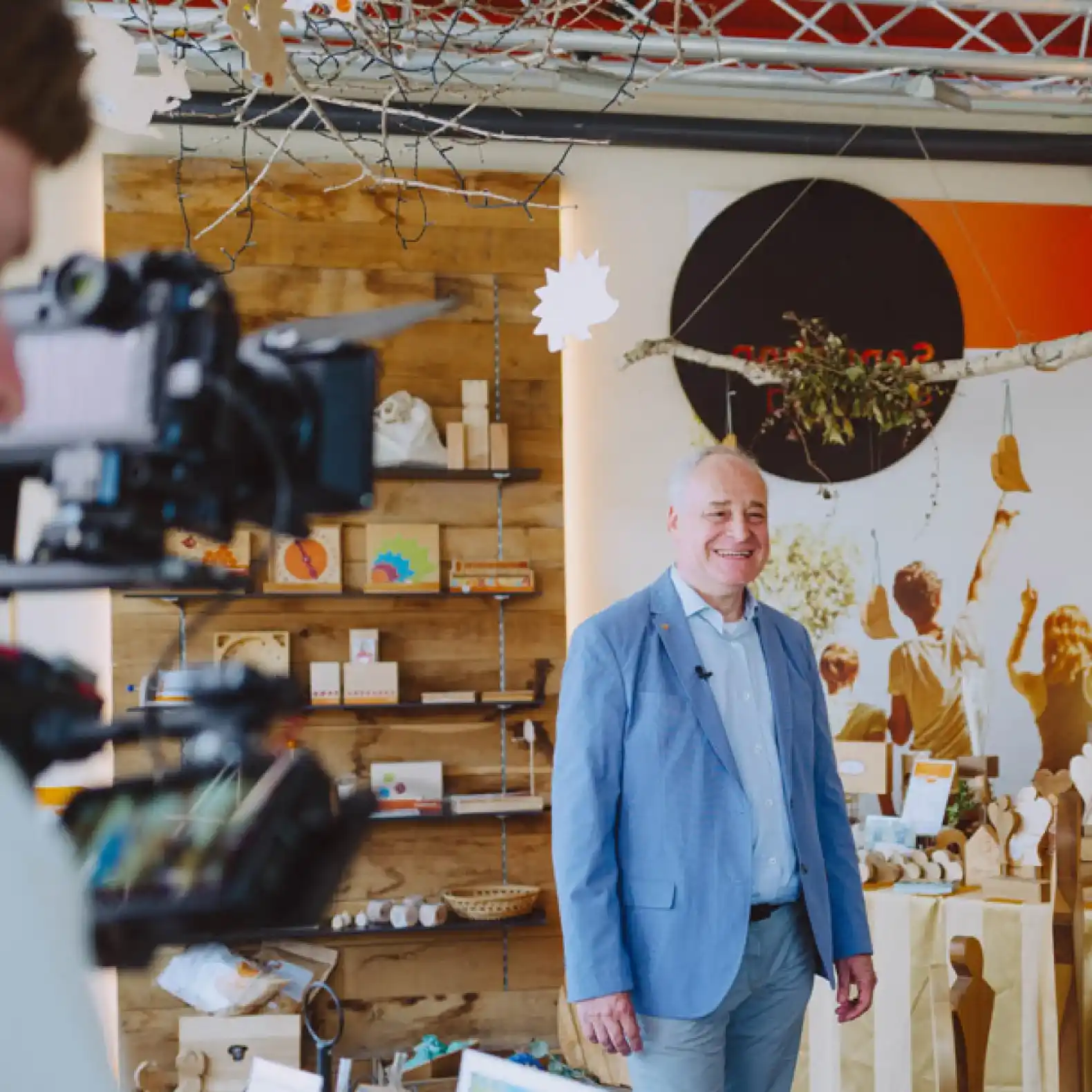 A man in a blue jacket stands smiling in front of a camera in a decorative room with wooden shelves and handmade objects. In the background, there is colorful wall decoration and hanging branches.