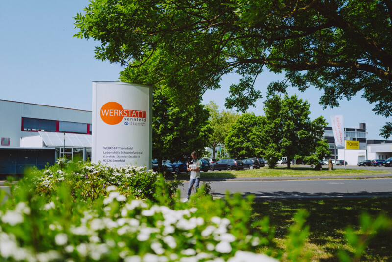 A sign reading "WERKSTATT Sennfeld" stands in front of a building. Trees and parked cars can be seen in the background, while sunny weather illuminates the scene.