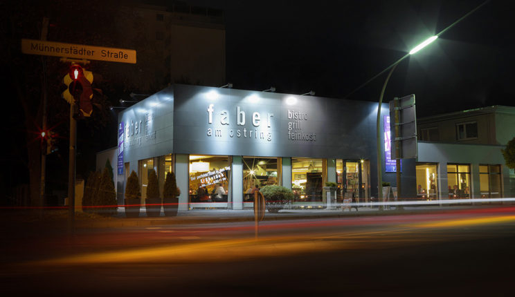 A nighttime shot of a restaurant with the illuminated sign "Faber am Ring" on the façade. In front of the building, the lights of passing cars are blurred. A traffic light and a street sign with the inscription "Münnersdörfer Straße" are located on the left side of the image.