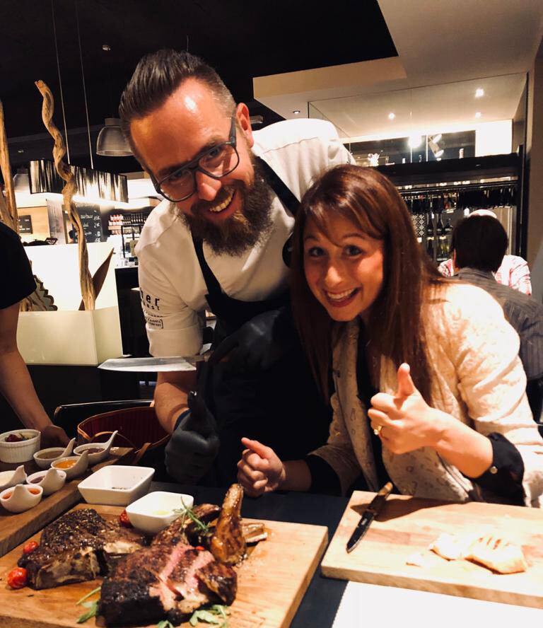 A chef and a woman smile at the camera, with grilled meat on a cutting board in front of them.