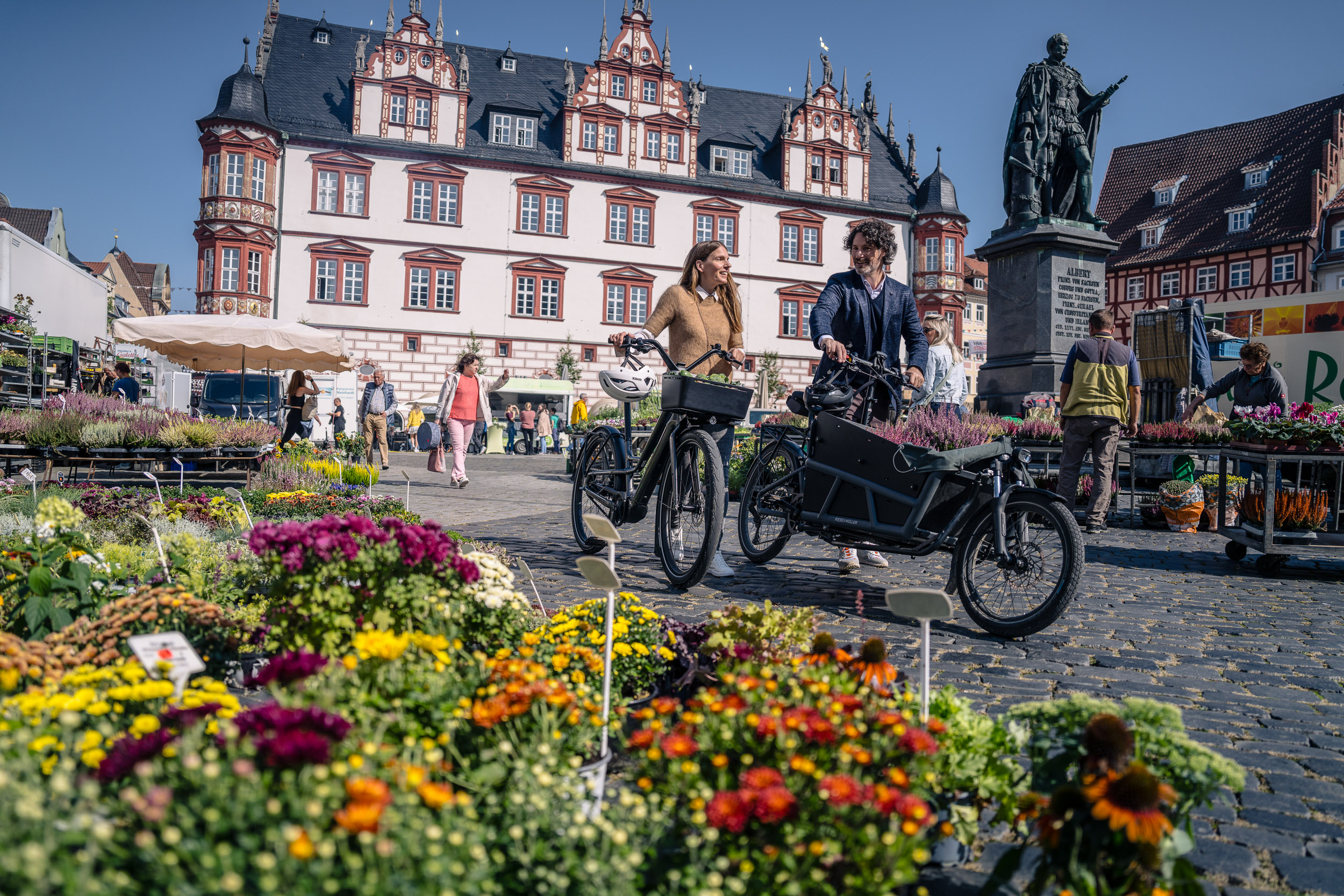 Two people with bikes stroll through a colorful flower market against a historic backdrop.