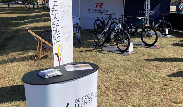 An exhibition stand for the Deutsche company bike with an advertising table and an outdoor display, surrounded by bikes and tents from various brands.