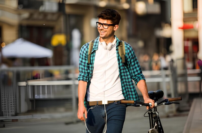 A young man wearing glasses, headphones, and a backpack pushes his bike through a busy city street and smiles.