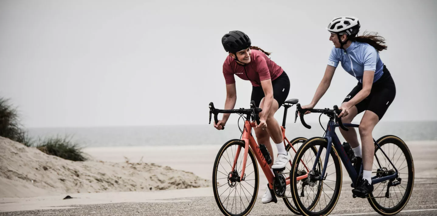 Two women are riding racing bikes on a sandy path near the coast. Both are wearing bike helmets and athletic clothing. The woman on the left is riding an orange bike, and the woman on the right is riding a blue bike. They smile at each other as they ride side by side.